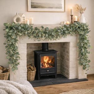 Può includere: Una scena accogliente con camino, caratterizzata da una stufa a legna nera con fuoco acceso. Il camino è decorato con una ghirlanda di foglie verdi e fiori bianchi, candele e vasi decorativi. Cesti di legno e altri elementi decorativi sono nelle vicinanze.