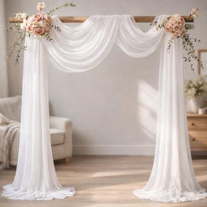 May include: A white wedding arch with draped fabric and floral arrangements. The arch is made of a wooden beam supported by two vertical posts. Soft white fabric is draped over the beam. Floral arrangements of pink and cream-colored roses and greenery adorn the top.
