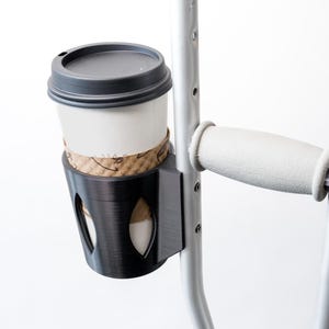 May include: A black 3D-printed cup holder attached to a silver crutch. The holder has a circular opening for a coffee cup with a black lid. The crutch has a gray handle and a wing nut for adjustments. The cup is white with a brown paper sleeve.