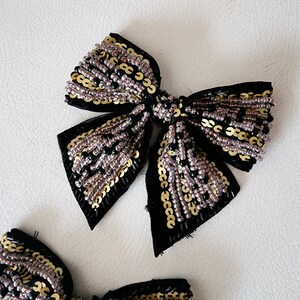 May include: Two black fabric bows with gold and silver sequins. The bows are tied together in the center.