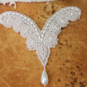 May include: A white beaded embellishment with a teardrop-shaped pearl pendant. The embellishment is in the shape of a V with two wings.