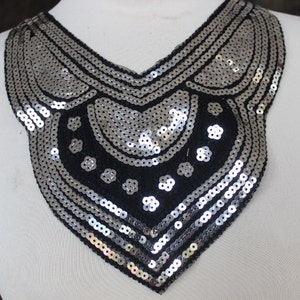 May include: A decorative silver sequin collar with a black base, displayed on a white mannequin. The collar features a V-neck design with intricate patterns of sequins and small floral accents. The sequins are arranged in curved and linear rows.