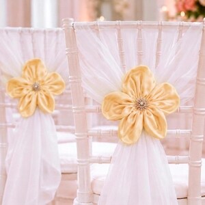 May include: A white wooden chair featuring a vertical spindle backrest draped with sheer white fabric. A large, pale yellow fabric flower with a central cluster of clear crystals is attached to the center of the chair back over the fabric.