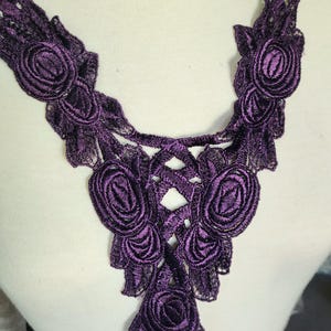 May include: A deep purple lace applique featuring a floral design. The applique has a series of embroidered rose-like flowers and intricate openwork details. This decorative trim is suitable for embellishing garments or craft projects.
