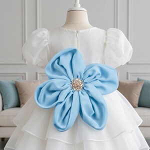 Puede incluir: Un vestido blanco con mangas cortas abullonadas y falda de capas. Una gran flor de tela azul claro con un centro de cristales transparentes y perlas está sujeta en la parte posterior de la cintura. El vestido tiene un cuello redondo.