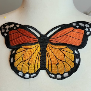 May include: Embroidered butterfly patch with orange, yellow, and black details. The butterfly has a black outline and detailed wing patterns. The patch is likely for clothing or accessories.