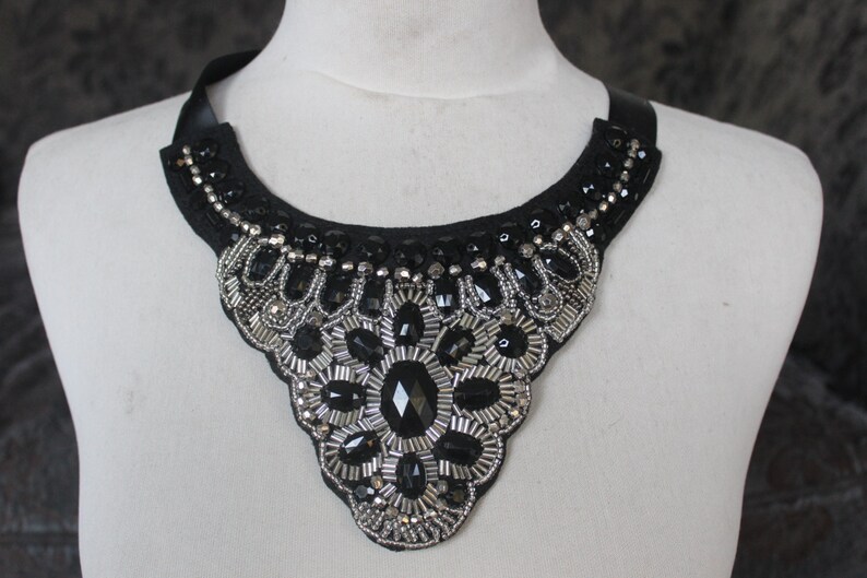 May include: A black choker necklace with silver and black bead embellishments. The necklace is in the shape of a V and has a large, central, black gemstone.