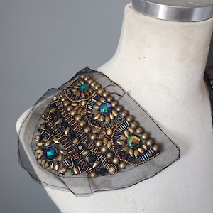 May include: An ornate shoulder embellishment featuring a black mesh base adorned with gold beads, iridescent jewels, and black seed beads. The design is triangular and is displayed on a white mannequin.