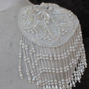 May include: A white beaded shoulder piece with a circular design and a fringe of pearls.