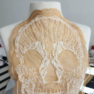 May include: A beige lace collar with an intricate floral design. The collar is made of delicate, sheer fabric and is displayed on a white mannequin. The lace pattern is white and detailed, creating a vintage aesthetic.