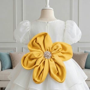 May include: A white dress featuring short puff sleeves and a tiered skirt. The back of the dress is adorned with a large, mustard yellow fabric flower centered with a cluster of clear, sparkling crystals, positioned at the waistline.