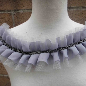 May include: A lavender-colored ruffled collar with a silver chain detail. The collar is made of layered fabric and tulle, creating a delicate and elegant look. This accessory could be used for cosplay or costume.