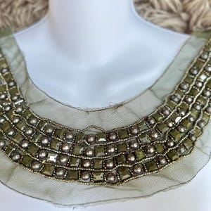 Puede incluir: Cuello con cuentas verde oliva con un delicado ribete de malla. El cuello presenta filas de cuentas cuadradas y redondas en plata y verde oliva, creando un acento decorativo en el escote.