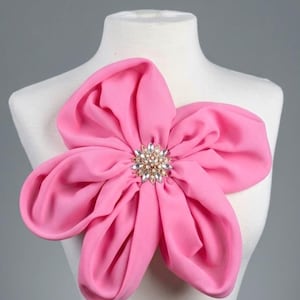 May include: A large, vibrant pink fabric flower, possibly a brooch or decorative accent, is displayed on a mannequin. The flower features multiple petals and a central embellishment with clear and gold-toned rhinestones. The background is a neutral gray.