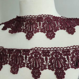 May include: Two strips of burgundy lace with a scalloped edge and an ornate floral design. The lace is displayed on a white mannequin head, showcasing the intricate details and rich color of the fabric. Perfect for sewing and craft projects.