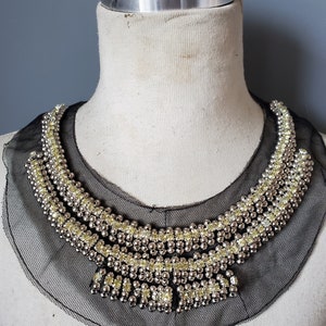 May include: A black mesh collar with a silver and yellow beaded embellishment. The collar is on a white mannequin.