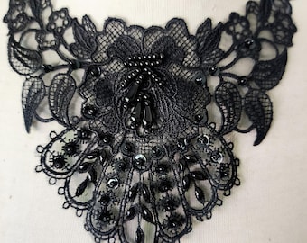 Puede incluir: Un cuello de encaje negro con intrincados patrones florales y de hojas. El cuello está adornado con cuentas y lentejuelas negras, creando un diseño delicado y ornamentado. El encaje se encuentra sobre un fondo blanco.
