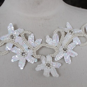 May include: A white floral applique with iridescent sequins and faux pearl accents. The design features multiple flower shapes with rhinestone centers, connected by a beaded vine-like structure. Two pins secure the applique to a mannequin.