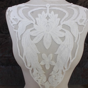 May include: White lace applique with a floral design. The applique is made of delicate lace and features a large flower in the center with smaller flowers and leaves around it.