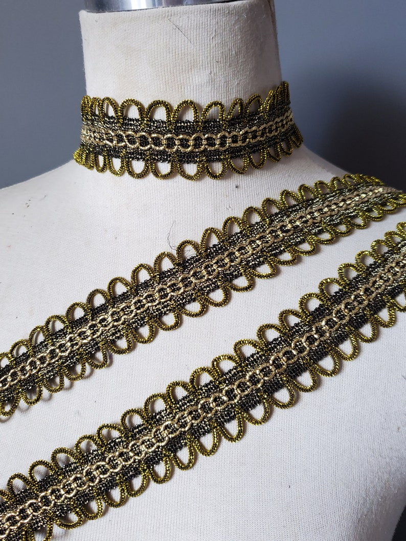 Puede incluir: Un adorno decorativo dorado y negro y un collar gargantilla a juego. El adorno y la gargantilla presentan un dise&ntilde;o de borde festoneado con un patr&oacute;n de cadena en el centro. El adorno se muestra en un maniqu&iacute;.