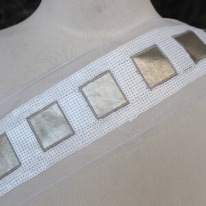 May include: White sheer fabric with a gold trim of square sequins. The trim is sewn on in a straight line.