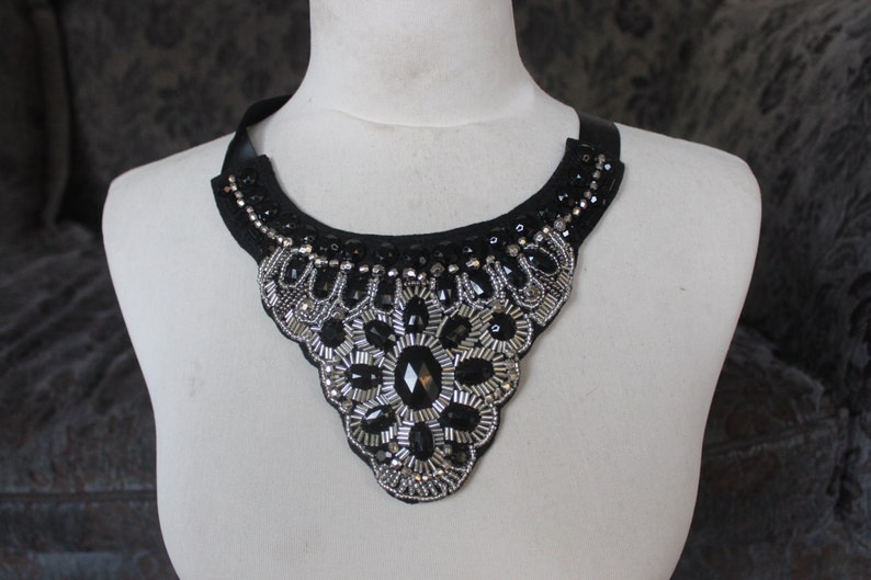May include: A black choker necklace with silver and black bead embellishments. The necklace is in the shape of a teardrop and has a scalloped edge.