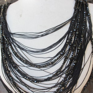 May include: A black beaded necklace with multiple strands of black thread. The necklace is layered and has a cascading effect.