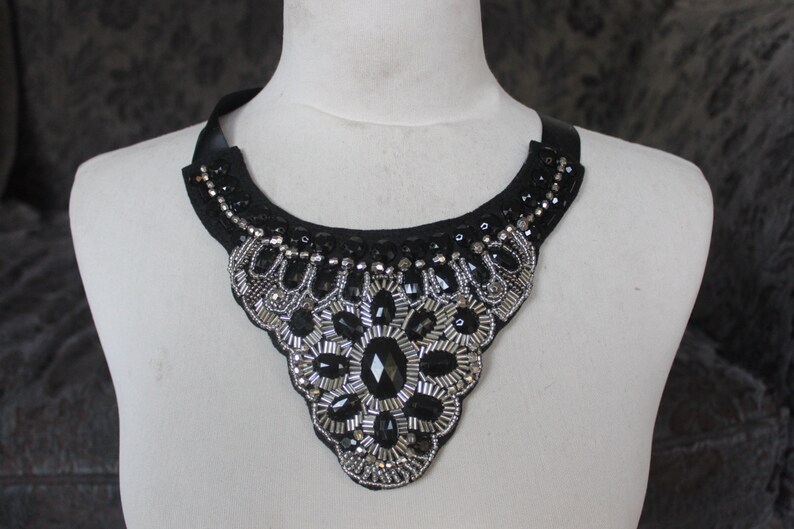 May include: A black and silver beaded statement necklace with a large, ornate design. The necklace is made of black fabric with silver beads and sequins.