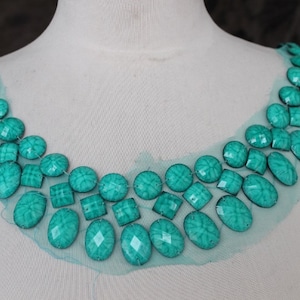 May include: Teal blue oval and square shaped rhinestones sewn onto a sheer turquoise fabric. The rhinestones are arranged in a curved pattern.