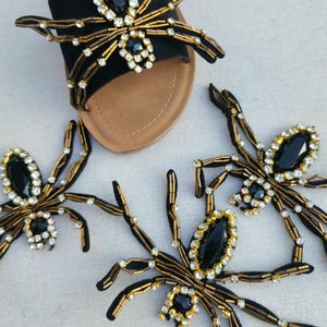 May include: Black slide sandals with spider decorations. Each spider has a black gem body, gold beaded legs, and clear rhinestone accents. The sandals have a tan sole.