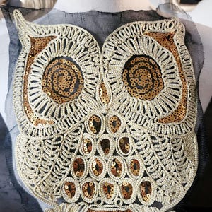 Op de afbeelding: Een gouden en witte uil applique met pailletten, op een zwart gaasstof. De uil is gedetailleerd met ingewikkelde stiksels en pailletten.
