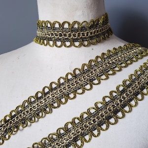 Puede incluir: Ribete decorativo dorado y negro y un collar gargantilla a juego. El ribete presenta un borde festoneado y un dise&ntilde;o en forma de cadena. El collar est&aacute; sobre un maniqu&iacute;. El ribete probablemente se utiliza para embellecer ropa o accesorios.