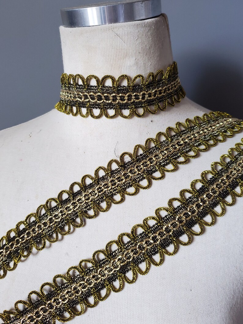 Puede incluir: Ribete decorativo dorado y negro y un collar gargantilla a juego. El ribete presenta un borde festoneado y un dise&ntilde;o en forma de cadena. El collar est&aacute; sobre un maniqu&iacute;. El ribete probablemente se utiliza para embellecer ropa o accesorios.