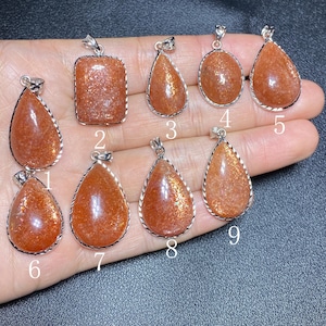 May include: A collection of nine sunstone pendants in various shapes, including teardrop and rectangular. Each pendant has a silver-colored bail and a shimmering, orange-brown hue. The pendants are displayed on a hand.
