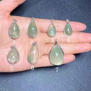 Puede incluir: Siete colgantes en forma de lágrima con engastes plateados. Los colgantes tienen un aspecto verde claro y translúcido con inclusiones internas. Cada colgante es único, con variaciones de color y patrón. Los colgantes se muestran en una mano.