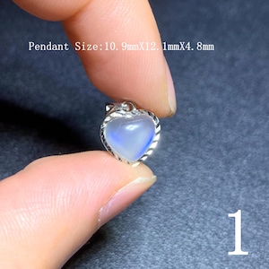 Puede incluir: Un colgante en forma de corazón con una piedra iridiscente azul y blanca, engastado en un marco de metal plateado. El colgante mide aproximadamente 10,9 mm x 12,1 mm x 4,8 mm. El número "1" es visible en la esquina inferior derecha.
