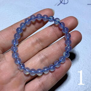 Puede incluir: Una pulsera de cuentas azules con cuentas redondas translúcidas. Las cuentas están ensartadas en un fino cordón elástico azul. La pulsera se muestra en la mano de una persona.
