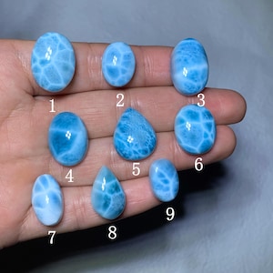 Puede incluir: Nueve piedras preciosas de larimar pulidas en varias formas, incluyendo ovaladas y en forma de lágrima. Las piedras muestran una gama de tonos azul claro con vetas blancas, creando un patrón similar a las olas. Las piedras preciosas están numeradas del 1 al 9.