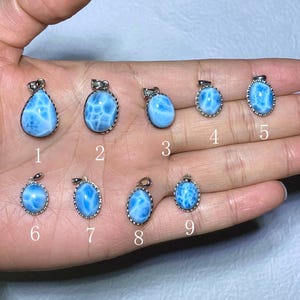 以下が含まれることがあります： 銀色の枠に収められた、9つのラリマーの宝石ペンダントのコレクション。ペンダントは、しずく型や楕円形など様々な形をしており、白のマーブル模様が入った水色の色合いです。各ペンダントには番号が振られています。