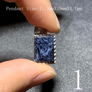 Puede incluir: Un colgante rectangular con una piedra azul oscuro y un marco de metal plateado. La piedra tiene un aspecto veteado. El colgante mide 1,13 cm x 1,9 cm x 0,41 cm. El número 1 es visible en la esquina inferior derecha.