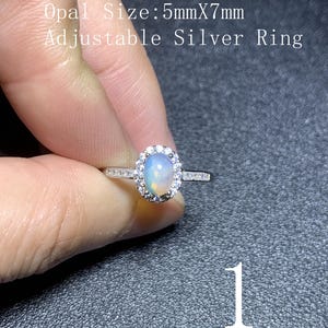 Puede incluir: Un anillo ajustable de plata con una piedra preciosa de ópalo ovalada, rodeada por un halo de piedras más pequeñas y transparentes. El ópalo mide 5 mm x 7 mm. El anillo es sostenido por una mano sobre un fondo oscuro. El texto en la imagen dice "Opal Size: 5mmX7mm Adjustable Silver Ring."