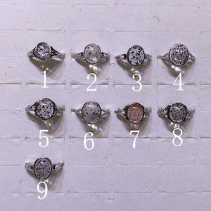 May include: Nine silver rings with oval-shaped gemstones. The gemstones vary in color and pattern, with some featuring speckled designs. The rings are displayed on a white surface, each labeled with a number from one to nine.