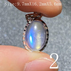 Puede incluir: Colgante ovalado con piedra lunar iridiscente engastada en un marco de metal plateado. La piedra mide 9,7 mm x 16,2 mm x 5,6 mm. El número "2" es visible en la imagen.