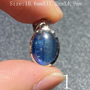 Puede incluir: Colgante ovalado de piedra preciosa azul con engaste de metal plateado. La piedra preciosa mide aproximadamente 10,6 mm x 17,2 mm x 4,9 mm. El colgante se sostiene entre los dedos sobre un fondo gris. El número "1" es visible.