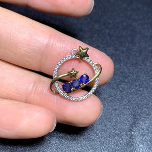 Puede incluir: Un colgante de temática celestial con un anillo dorado con una estrella y tres piedras preciosas azules. El anillo está incrustado con pequeñas piedras transparentes. Dos estrellas doradas están situadas encima y debajo del anillo.