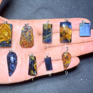 May include: A collection of nine gemstone pendants in various shapes, including rectangular, teardrop, and irregular forms. The pendants feature a mix of blue, gold, and brown colors, with silver-colored metal bails for hanging. The gemstones are displayed on a hand.
