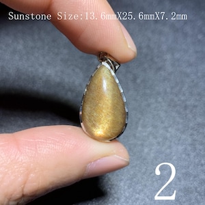Op de afbeelding: Een druppelvormige zonnesteen hanger met een zilverkleurige rand. De zonnesteen heeft een goudbruine kleur met een glinsterend effect. De tekst "Sunstone Size: 13.6mmX25.6mmX7.2mm" is zichtbaar. Afmetingen: 2,56 cm x 1,36 cm x 0,72 cm. Het nummer "2" is zichtbaar.