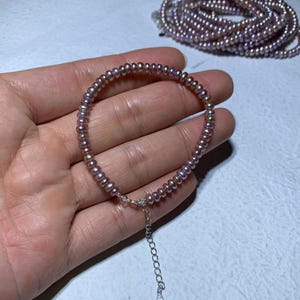Puede incluir: Una delicada pulsera con pequeñas perlas redondas de color malva ensartadas. La pulsera tiene un cierre plateado y una cadena ajustable. Una segunda pulsera similar es visible en el fondo, junto con una mano.