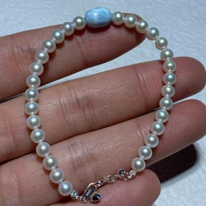May include: A delicate bracelet featuring small, round, white pearls strung together. A light blue, oval-shaped stone is centered on the bracelet. The clasp is silver and marked with "S925".