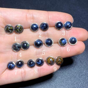Puede incluir: Una colección de nueve pares de pendientes de botón de piedras preciosas. Los pendientes presentan piedras redondas pulidas en varios tonos de azul, dorado y verde, engastadas en metal plateado. Las piedras tienen patrones y texturas únicos.